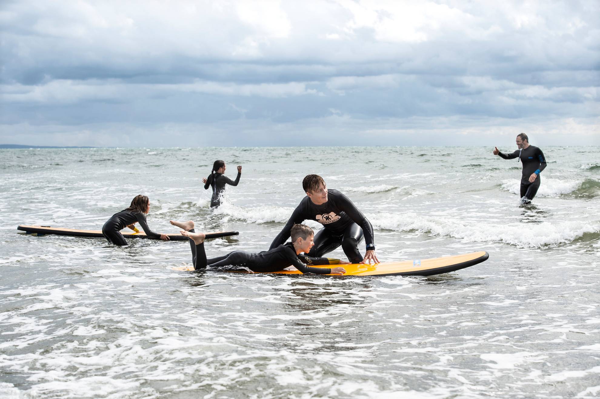 Två vuxna och tre barn står i vattenbrynet och två av barnen provar att surfa på varsin surfbräda.