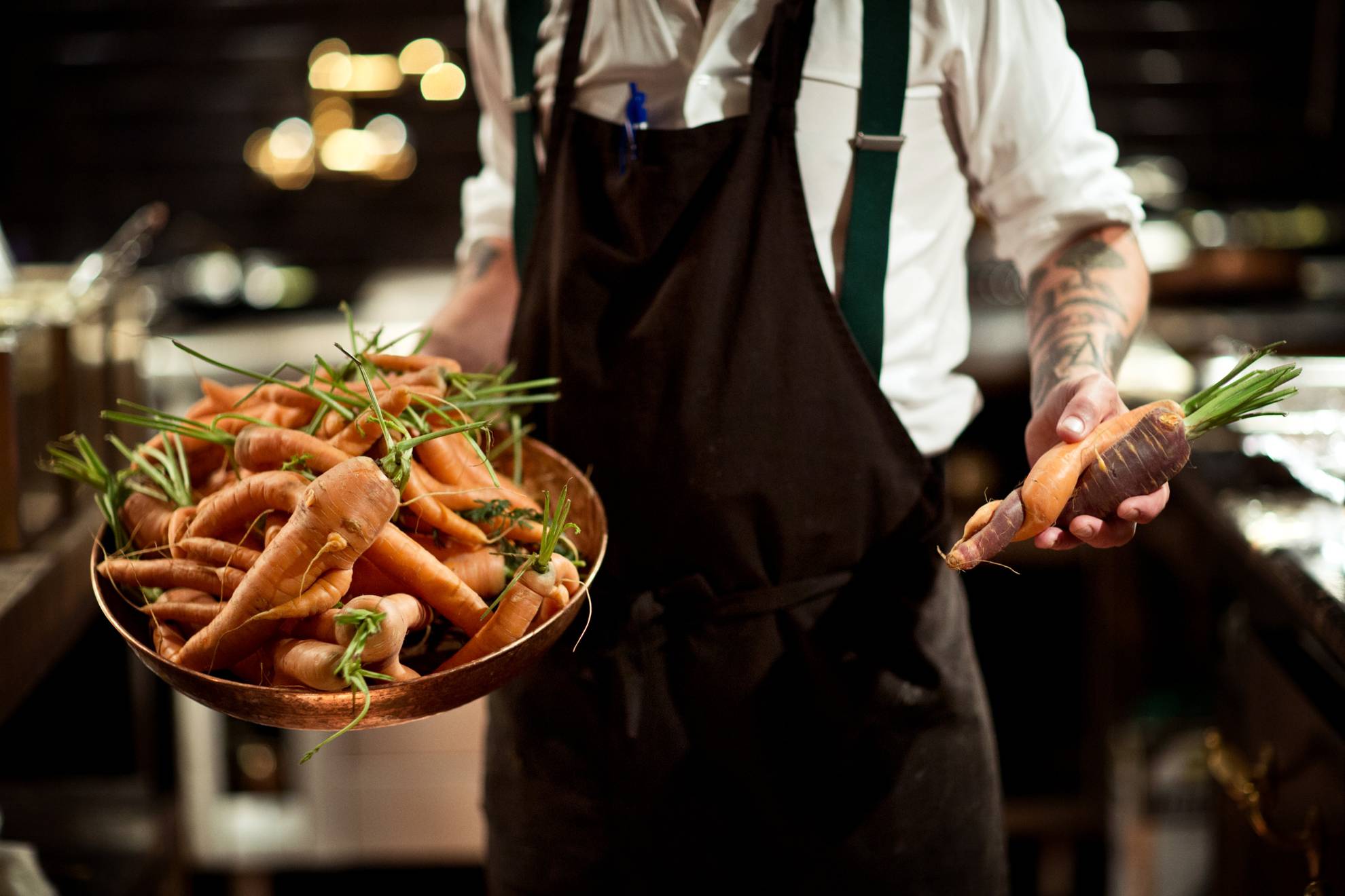 Bild från ett restaurangkök. En person håller fram en skål med morötter i olika former i ena handen och i andra handen fler morötter med blast.