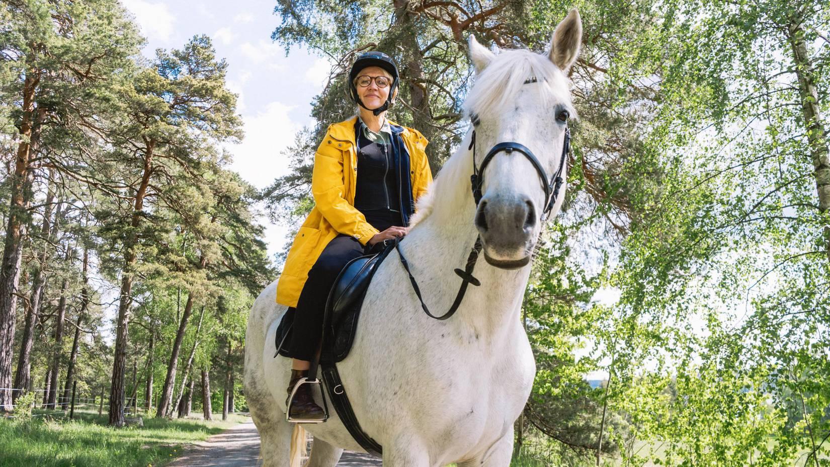 Kvinna i gul jacka sitter på en vit häst ute i skogen och ler mot kameran.