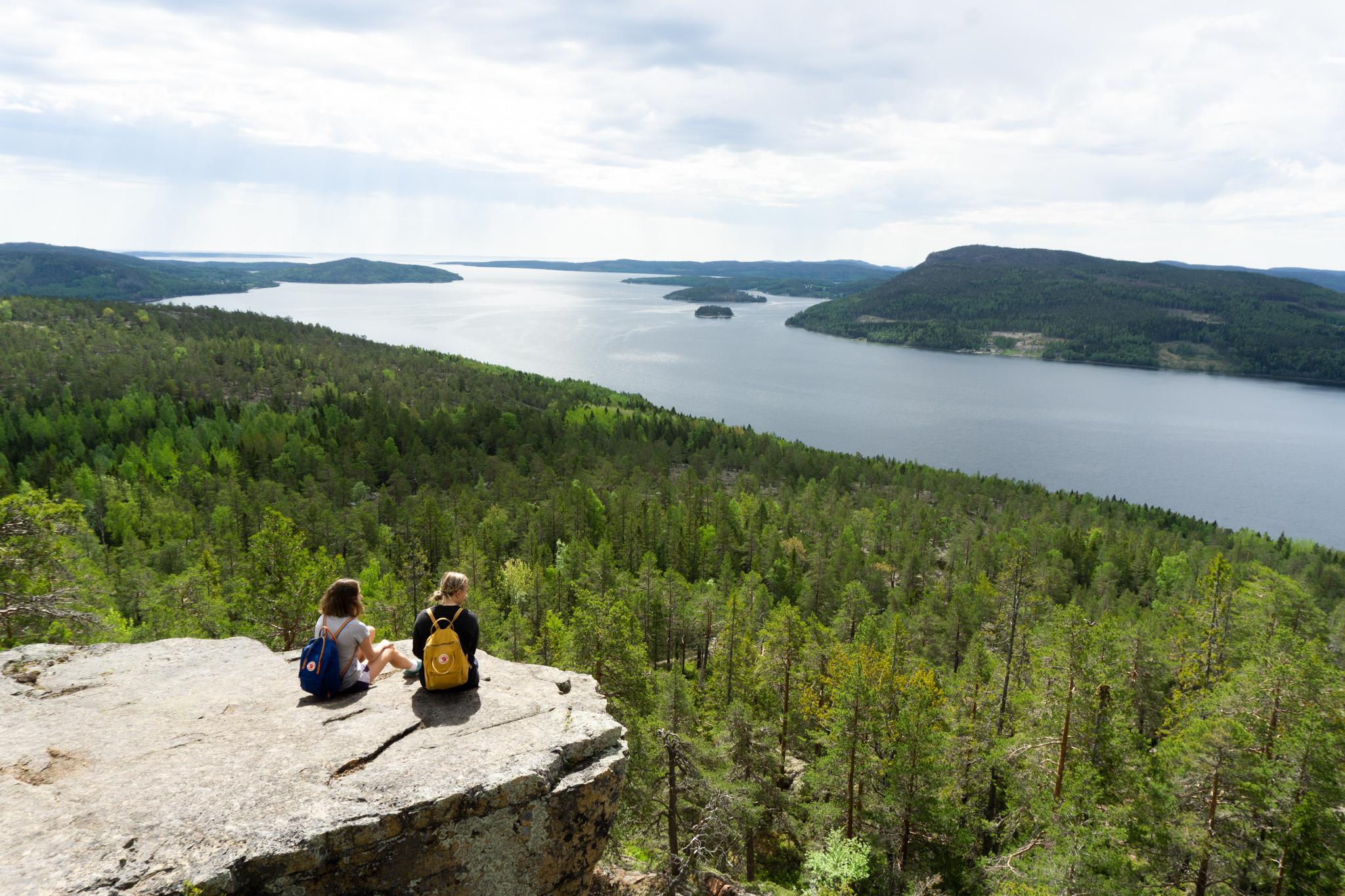 Två personer med ryggsäckar sitter på en klippavsats med utsikt över skog och en stor sjö.