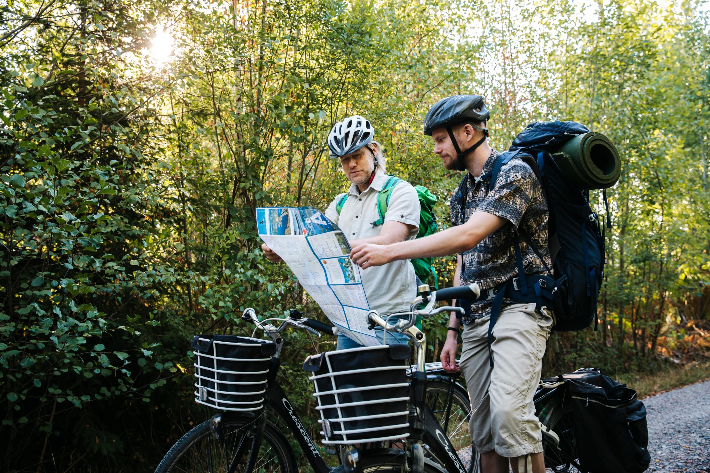 Två personer med ryggsäckar och cykelhjälmar står på en väg i skogen över sina cyklar och tittar på en karta.