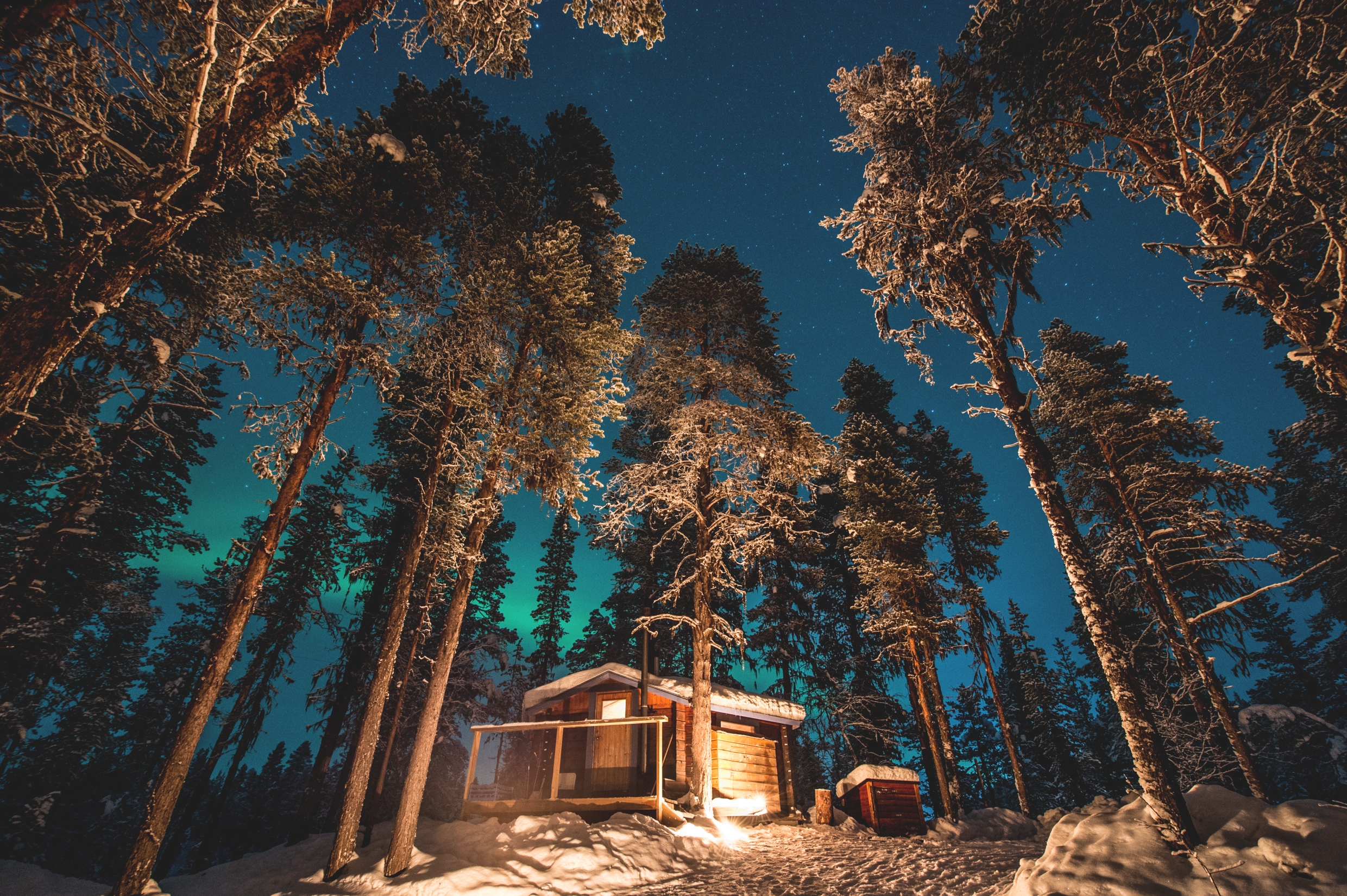 Under kvällstid, en snötäckt upplyst stuga i i skogen med höga träd runtomkring och norrsken i bakgrunden.