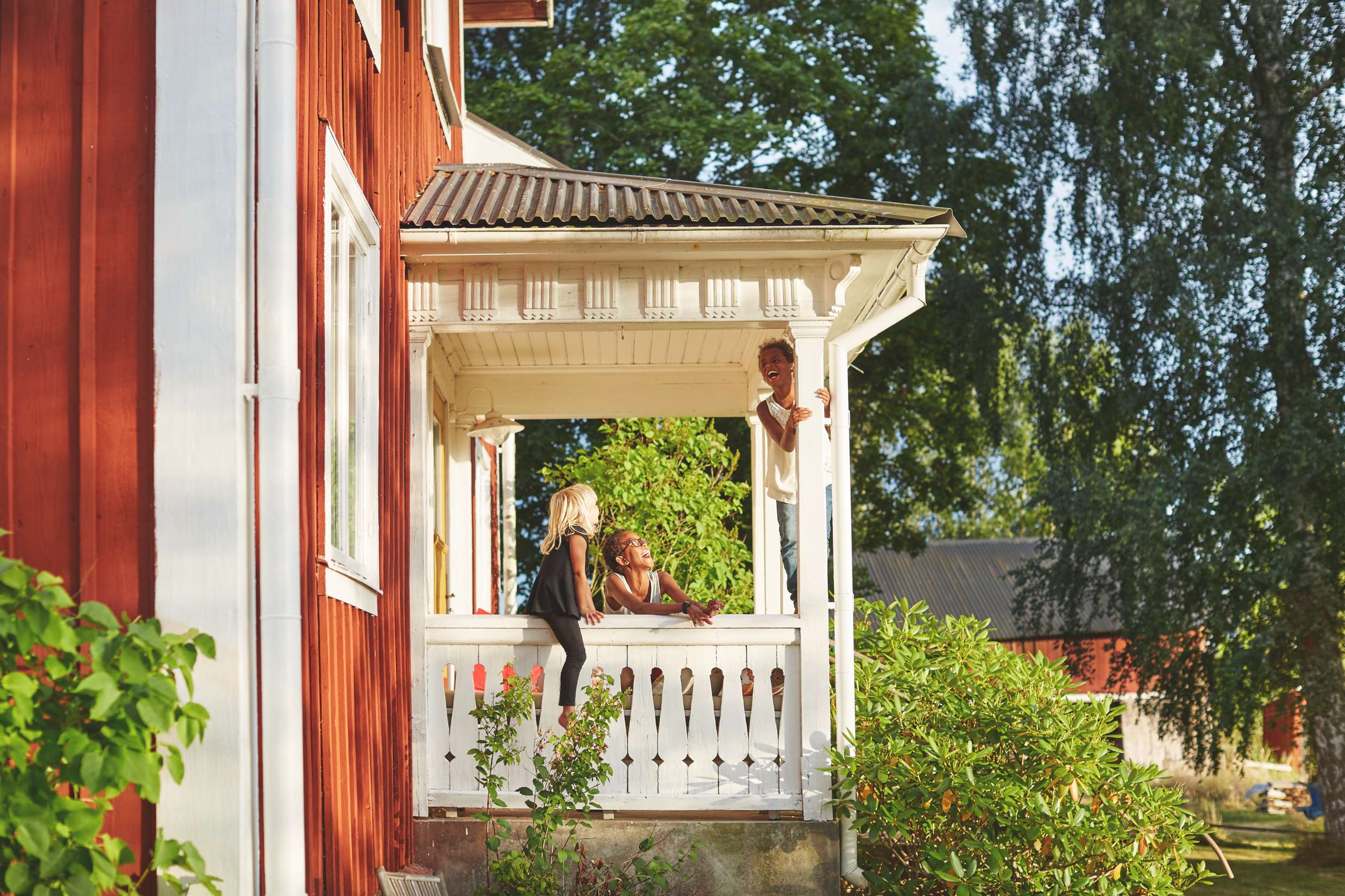 Tre barn leker på verandan till ett rött hus med vita knutar. Träden omkring blommar och solen lyser.