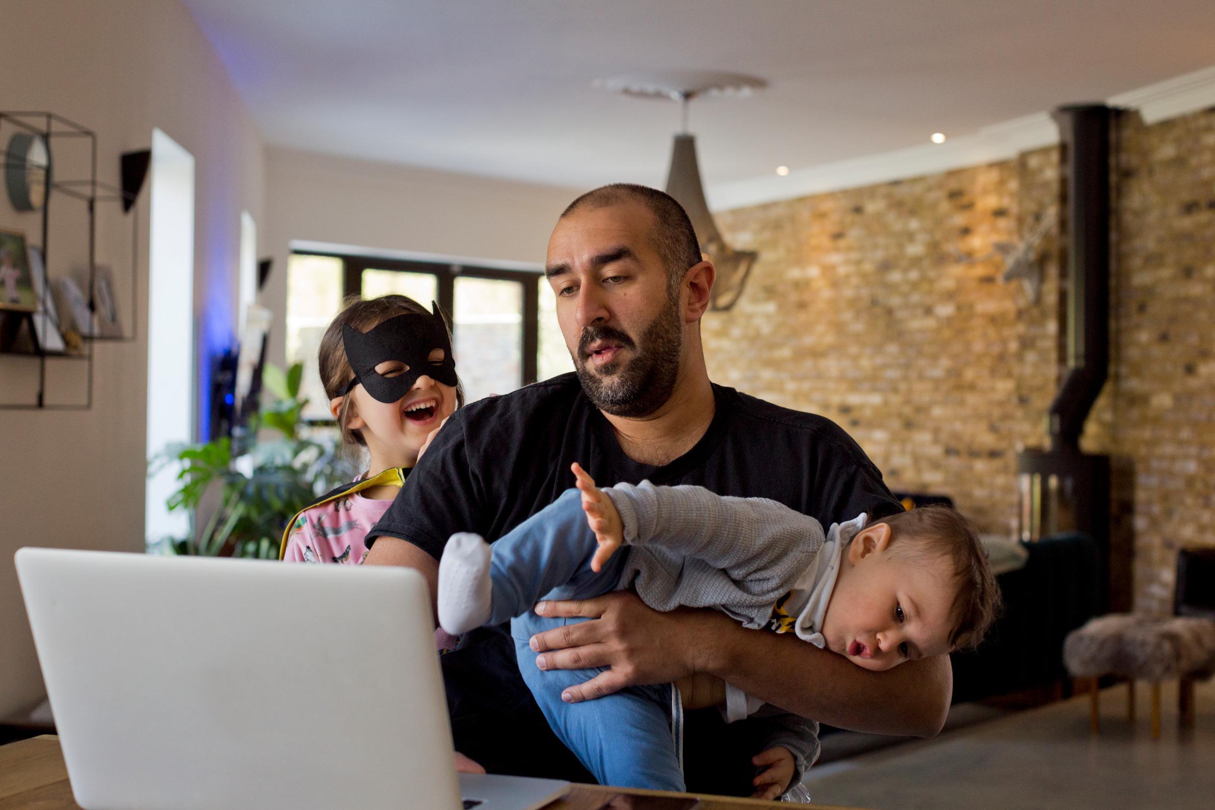 En man står inomhus bakom en dataskärm och håller en bebis på armen samtidigt som ett barn bakom honom pockar på uppmärksamhet.