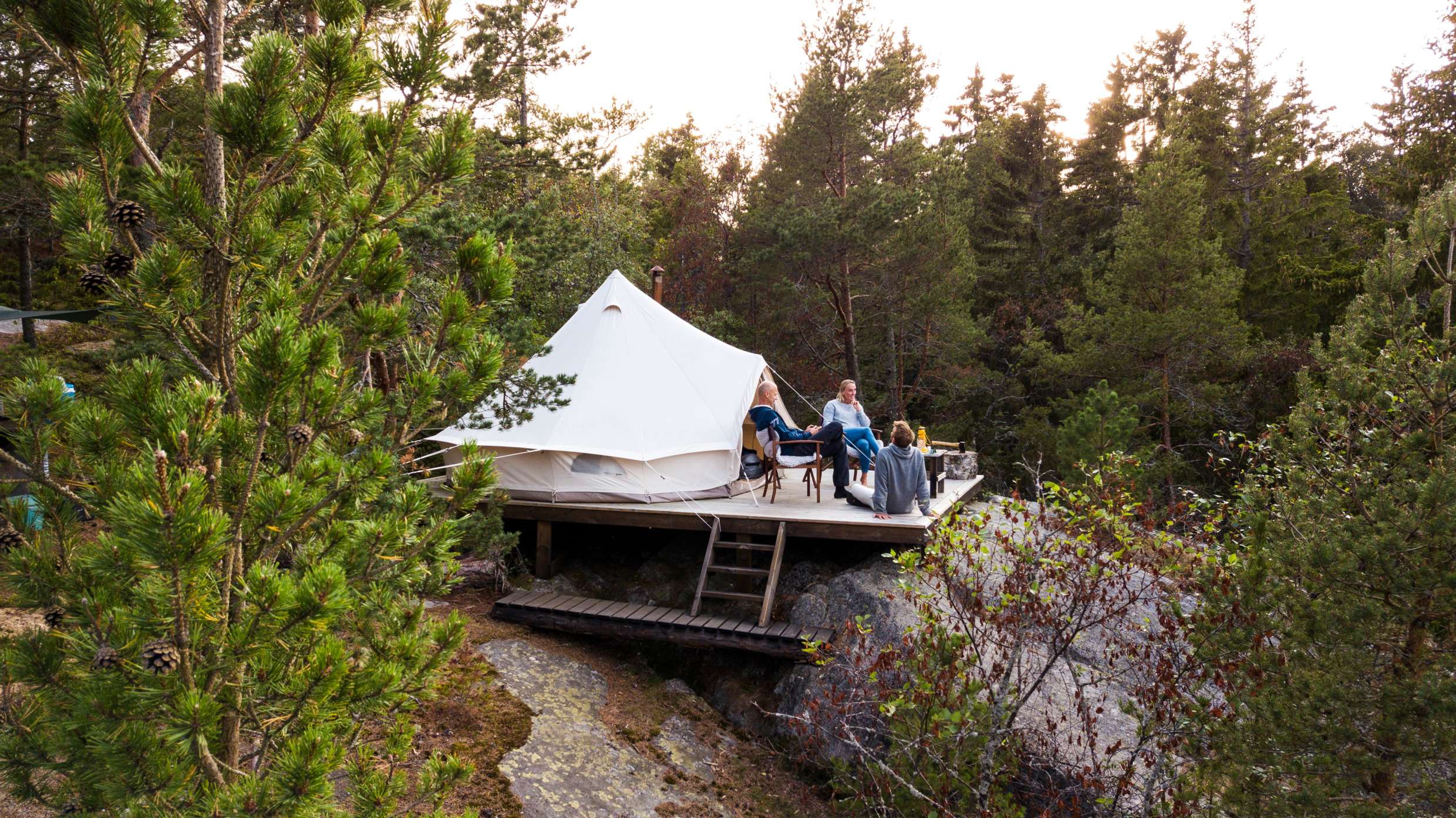 Ett stort tält på en träavsats på ett berg mitt i skogen vid en sjö. Framför tältet på trädäcket sitter flera personer.