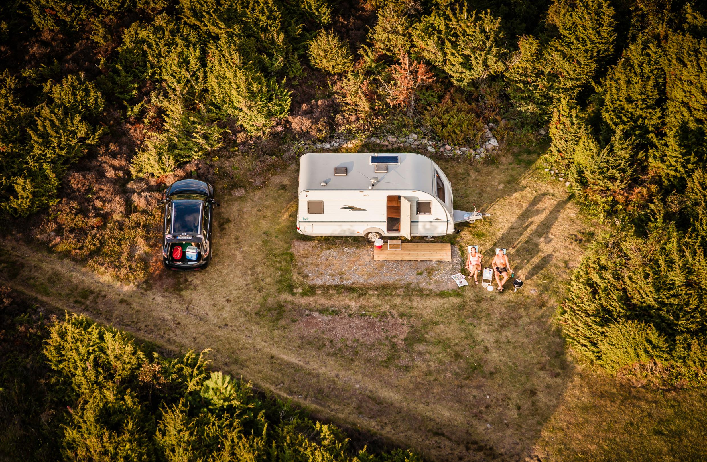 Bilden visar en husvagn uppställd på en naturnära campingplats. Två personer sitter i solstolar utanför och njuter i solen.