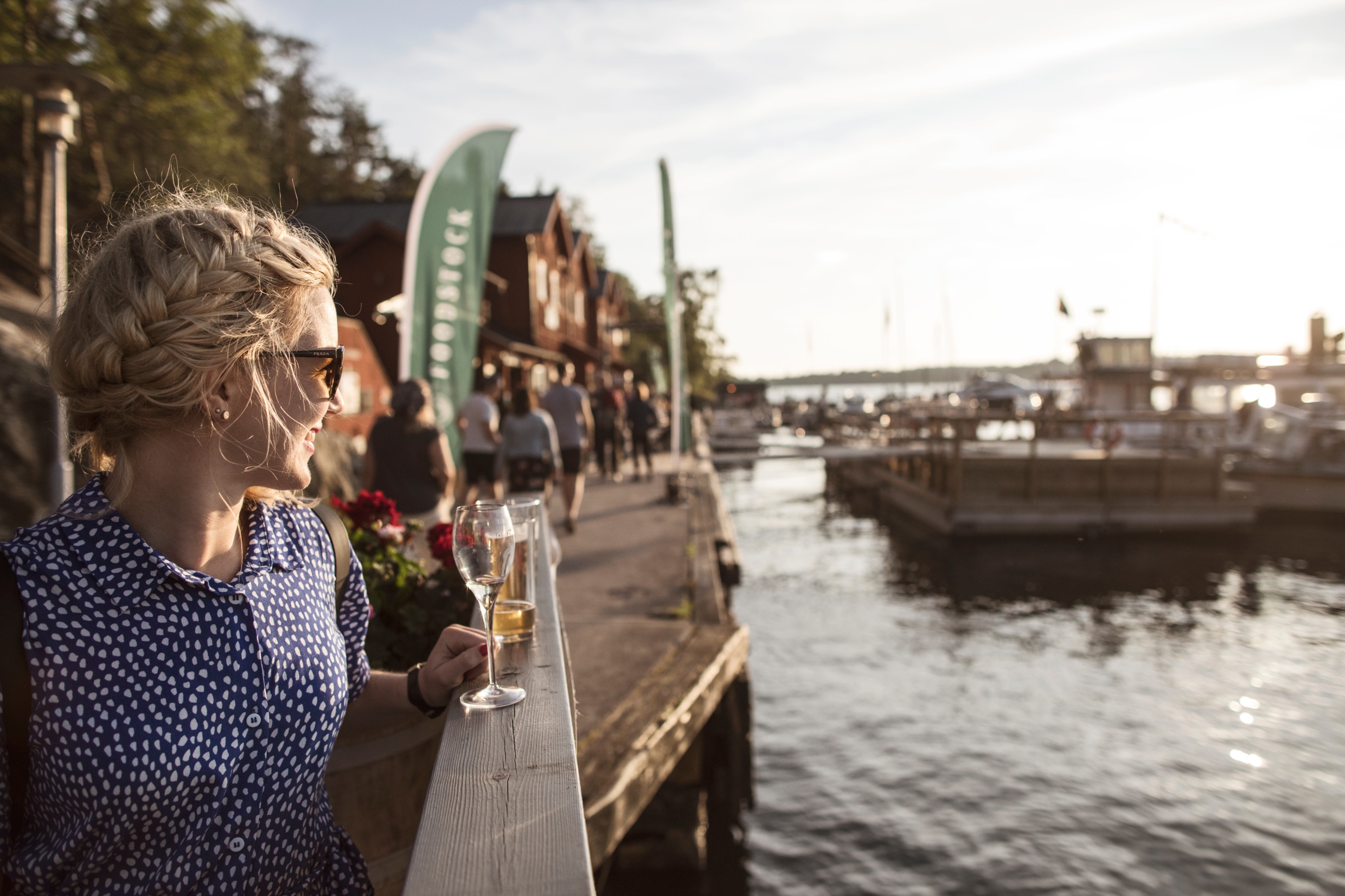 En sommarkväll i skärgården. En kvinna i en blommig klänning står på en kaj och blickar ut över hamnen i Fjäderholmarna med ett glas i handen.